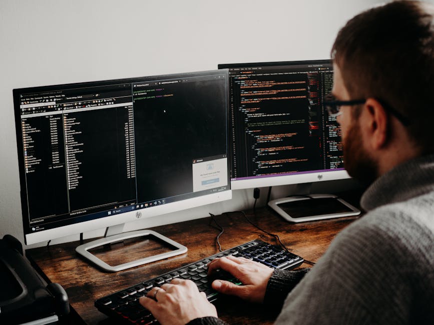 A person with short brown hair and glasses is working at a wooden desk with two large computer monitors displaying lines of code, one with a dark theme and the other with a lighter background. The wor