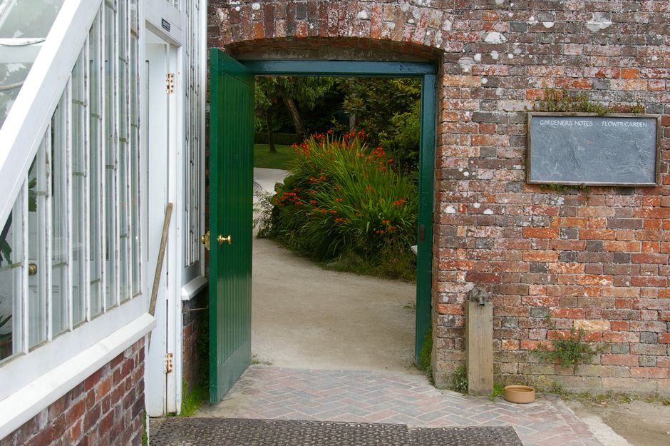 A small garden entrance framed by a red brick wall and a white-painted greenhouse with a metal grate window on the left. The open green gate reveals a pathway leading into a lush garden filled with vi