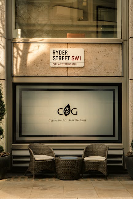 The image depicts a cozy outdoor seating area set against a building façade with large windows. In front of the window, there are two woven rattan chairs with light-colored cushions and a matching ro